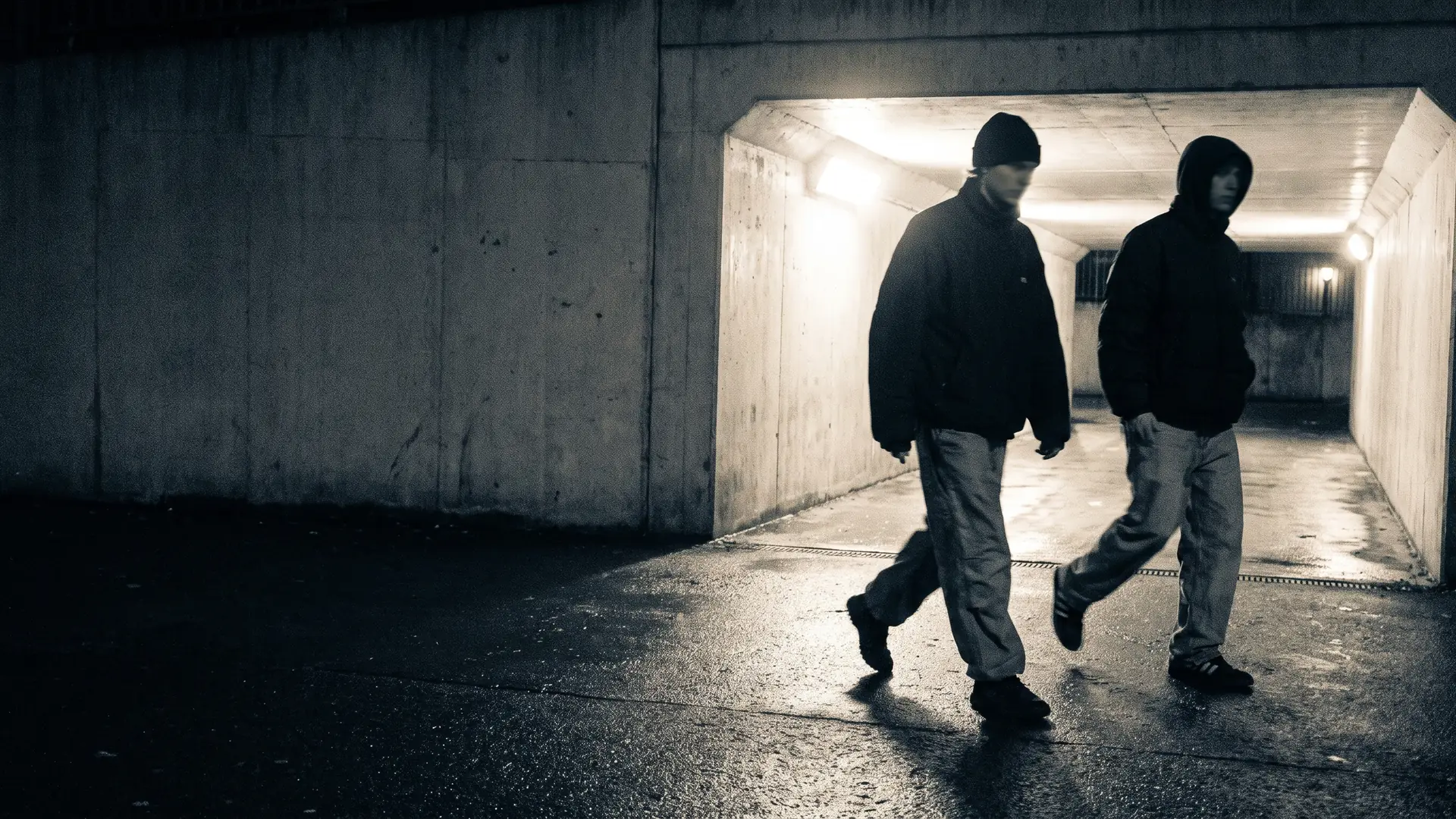 Two figures in streetwear walking at night through a lit concrete pedestrian underpass, wet asphalt underfoot